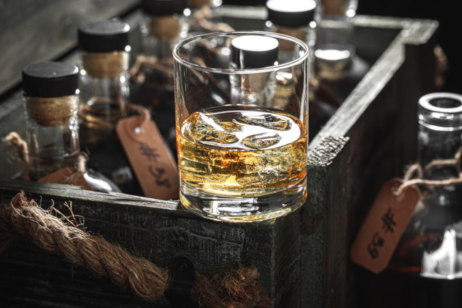 Whiskyglas mit Eis vor Flaschen in einer Holzkiste.