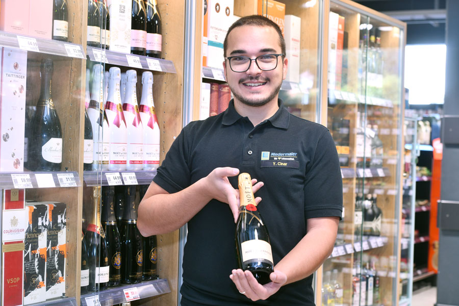 Mitarbeiter im Getränkeregal präsentiert eine Flasche Sekt vor Wein- und Spirituosenregal.